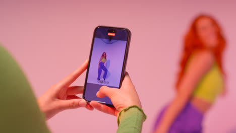 Studio-Shot-Of-Woman-Taking-Photo-Of-Friend-Dancing-On-Mobile-Phone-Against-Pink-Background-5